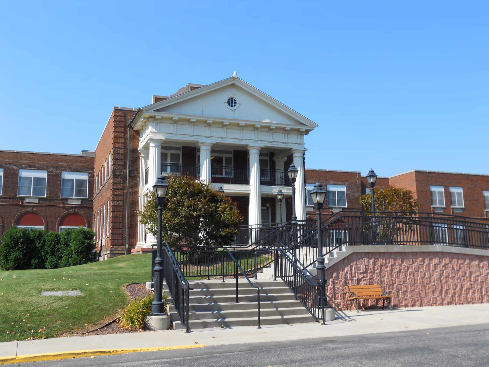 Front exterior view of Pleasant Ridge Manor, a large brick building with white columns and a triangular pediment above the entrance. There are stairs and a ramp leading up to the entrance, black metal railings, several lamp posts, a bench, and some trees and shrubs on a grassy slope.