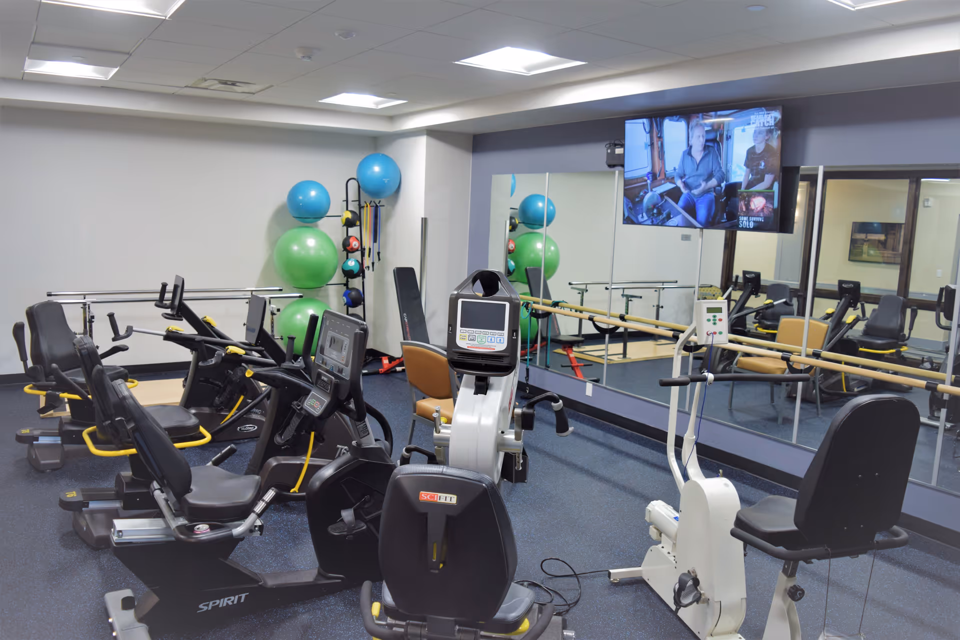 A fitness room with several exercise bikes and a rowing machine. There are large green and blue exercise balls stacked in the corner, a wall-mounted TV showing a program, and a large mirror covering one wall. The room has a blue carpet and white walls with ceiling lights.