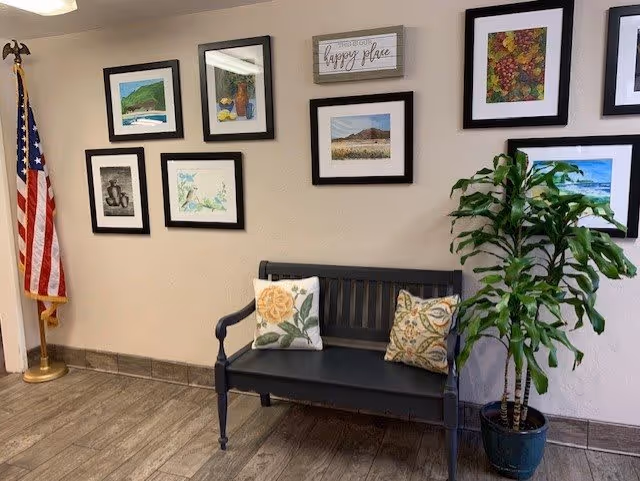 An interior corner of a room featuring a black wooden bench with two decorative pillows, a potted green plant to the right, and an American flag on a stand to the left. The beige wall behind the bench is decorated with several framed pictures and a sign that reads 'This is our happy place.' The floor has wood-like tiles.