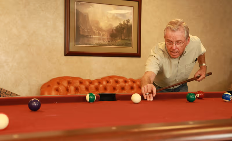 An elderly man playing pool on a red felt pool table in a room with a brown tufted leather couch and a framed landscape painting on the wall behind him.