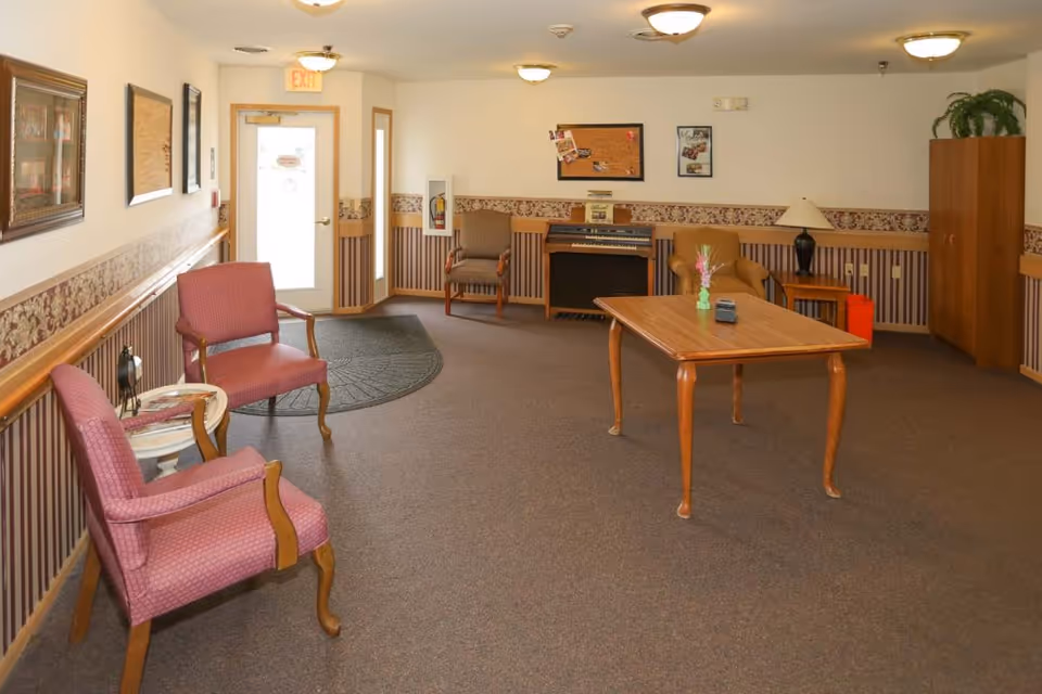 A common area in an assisted living facility featuring several upholstered chairs, a wooden table with a small vase of flowers, a piano against the far wall, framed pictures and bulletin boards on the walls, a lamp on a side table, and a cabinet with a potted plant on top. The room has carpeted flooring and soft lighting from ceiling fixtures.