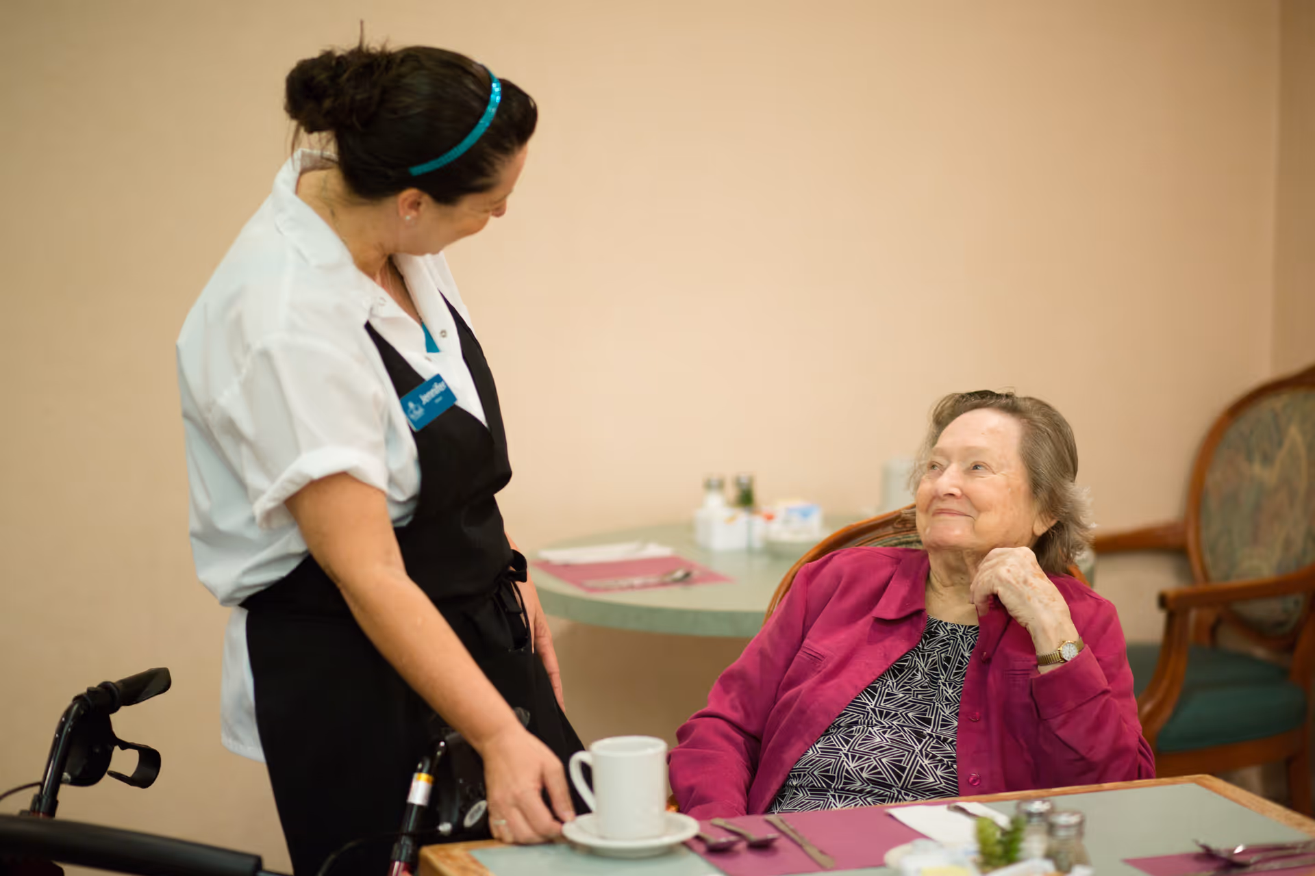 A caregiver talks with an elderly woman seated at a dining table in a dining room.