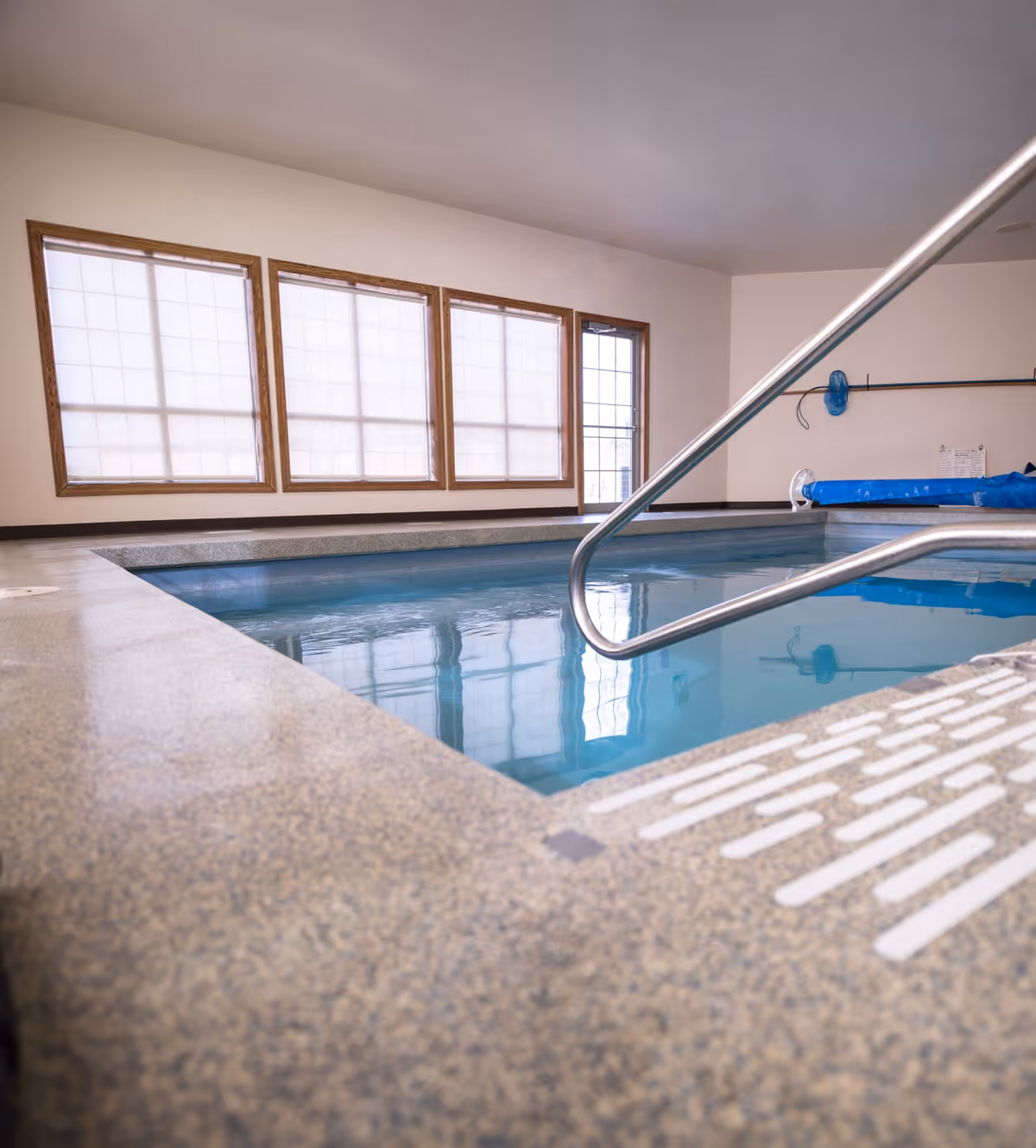 Indoor swimming pool with clear blue water, surrounded by a textured stone edge. Large windows with wooden frames and white blinds allow natural light into the room. A metal handrail extends into the pool for easy access.