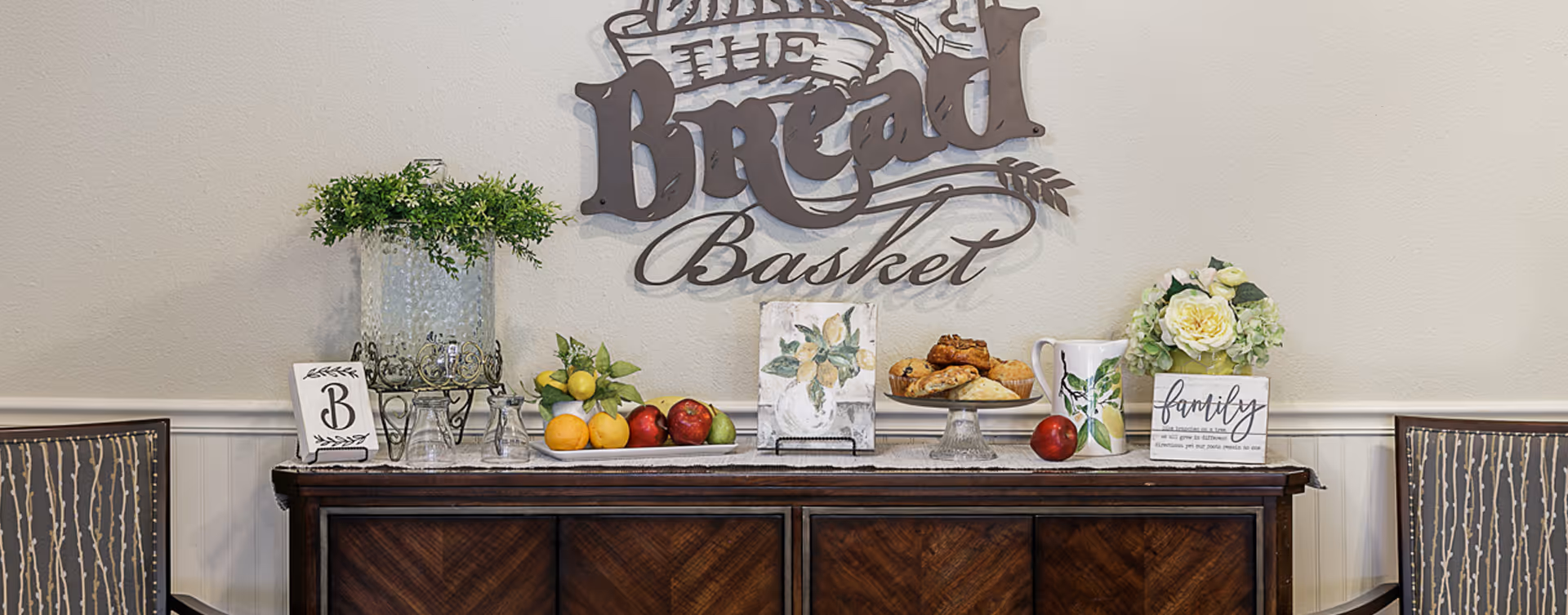 A wooden sideboard with decorative items including a glass water dispenser with greenery, a plate of assorted fruits, a plate of pastries, a pitcher, and floral decorations. Above the sideboard is a wall decoration that reads 'The Bread Basket'. Two chairs with patterned upholstery flank the sideboard.