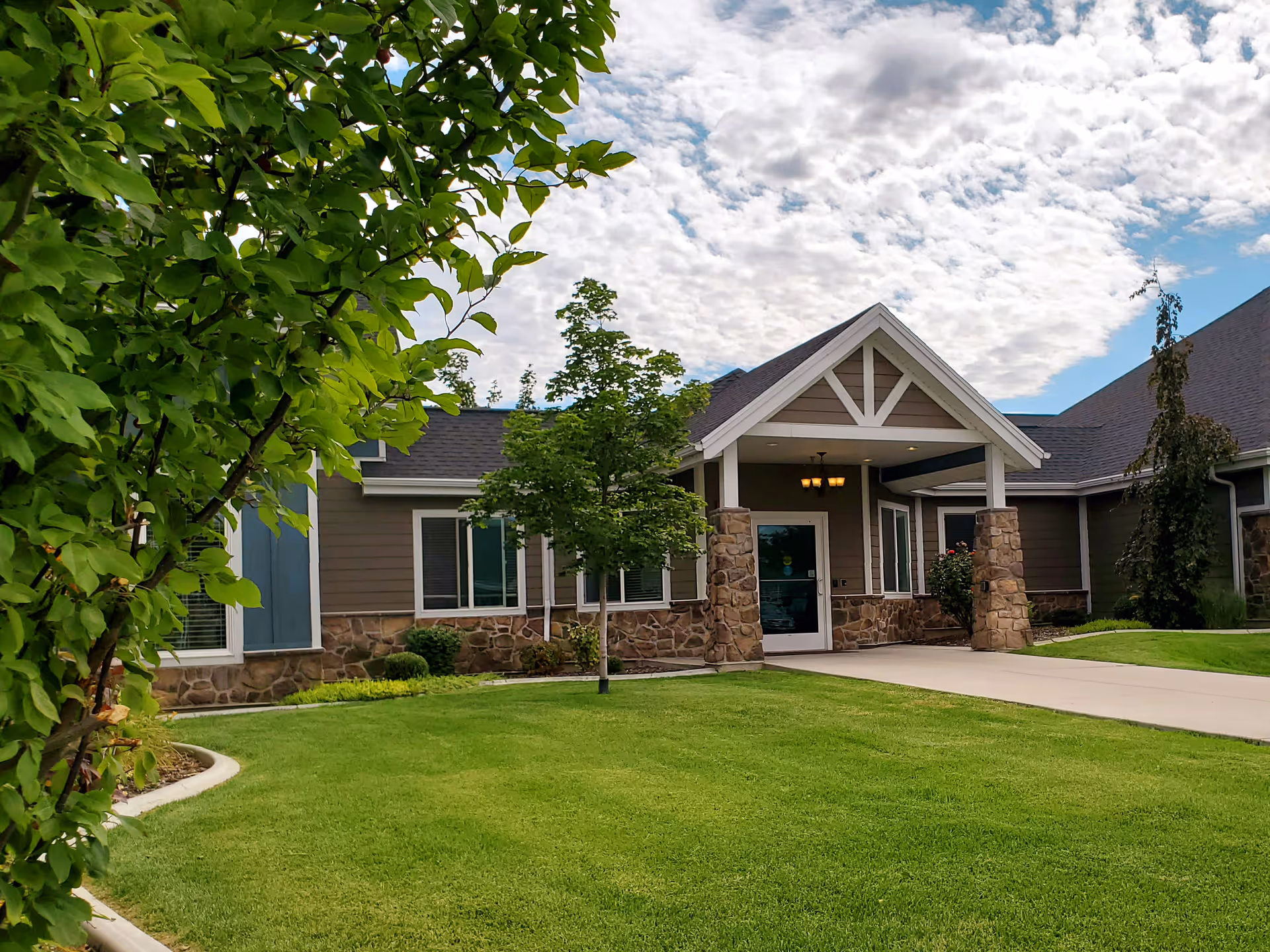 Exterior view of Cogir of Lotus Park facility showing a well-maintained lawn, trees, and a building entrance with stone pillars and a covered porch under a partly cloudy sky.