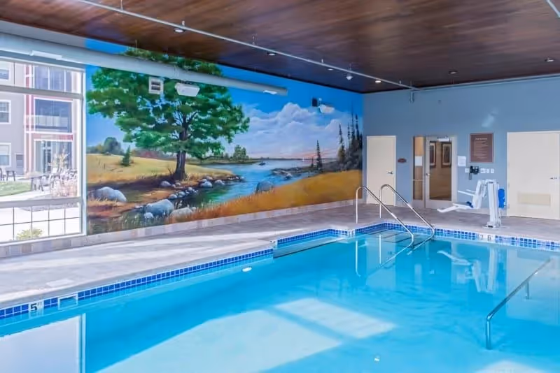Indoor swimming pool with clear blue water, metal handrails, and a mural on the wall depicting a scenic landscape with a tree, river, and sky. There are doors and a pool lift visible on the far side of the pool.