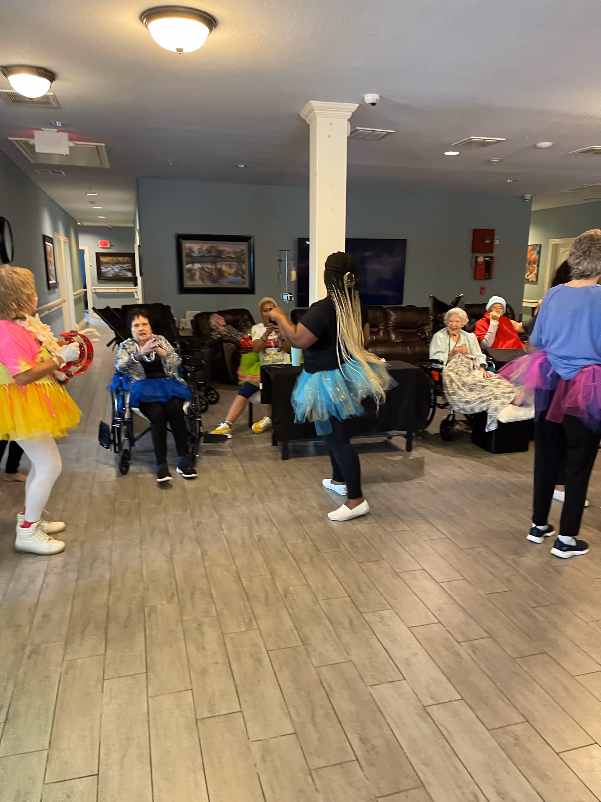 A group of elderly people and caregivers in a senior living facility common area. Some elderly residents are seated, including one in a wheelchair, while caregivers wearing colorful tutus and casual clothing engage with them. The room has wood-look flooring, a column in the center, and framed artwork on the walls.