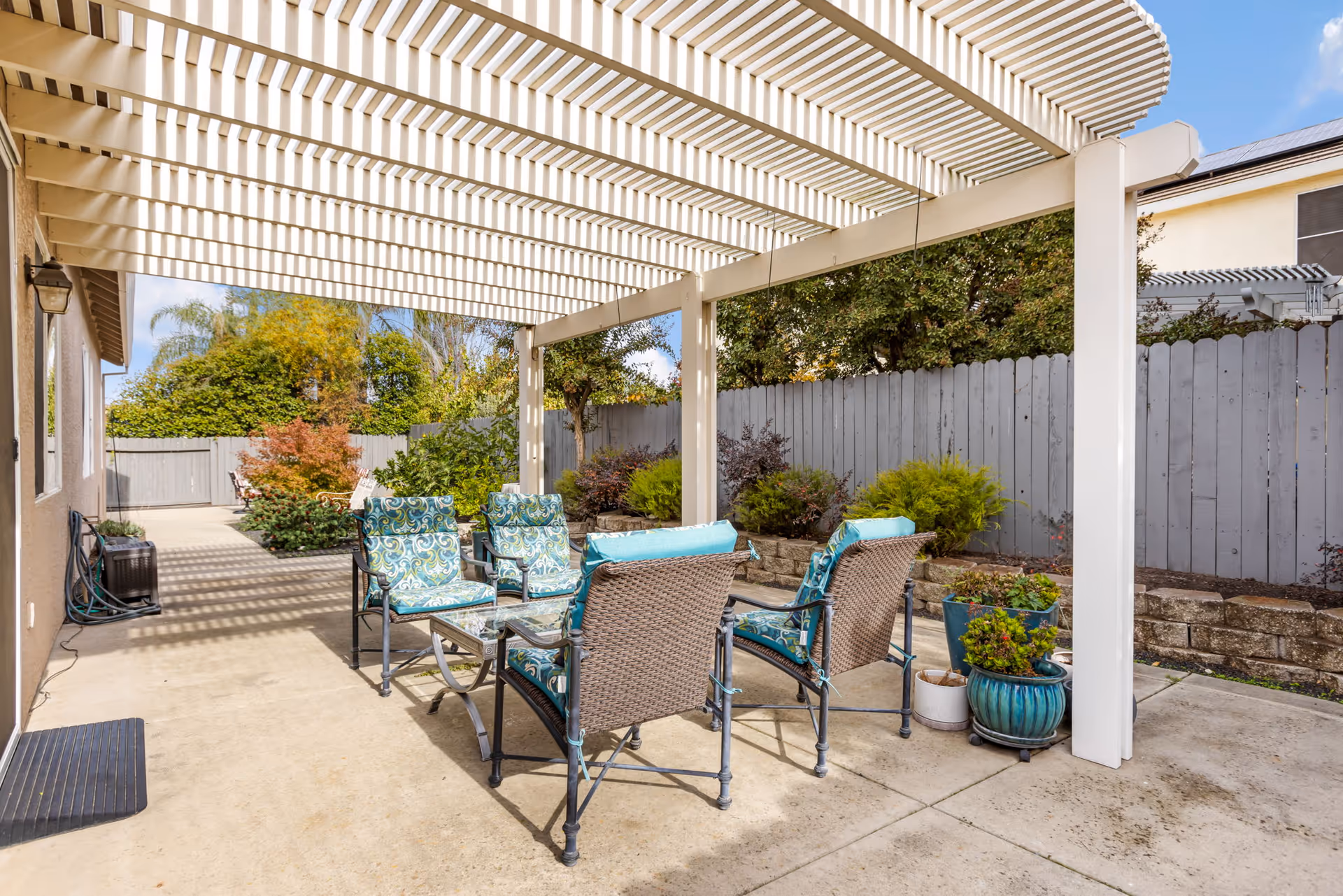 Outdoor patio area with a white pergola overhead casting striped shadows on the concrete floor. There are four cushioned chairs with blue and green patterned cushions arranged around a glass-top coffee table. The patio is bordered by a gray wooden fence and landscaped with various green shrubs and potted plants.