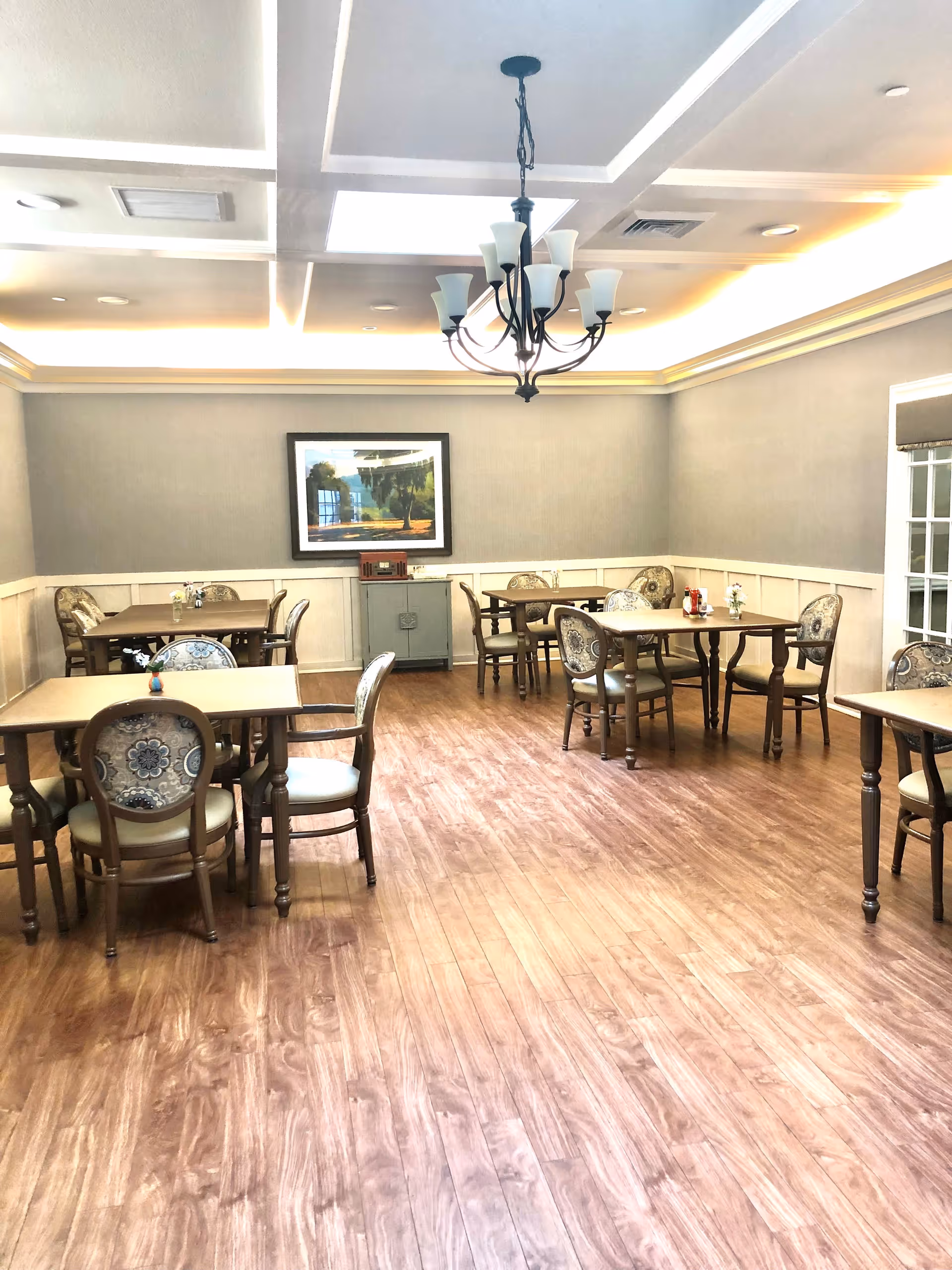 A bright dining room with wooden floors, multiple tables and upholstered chairs beneath a decorative chandelier.