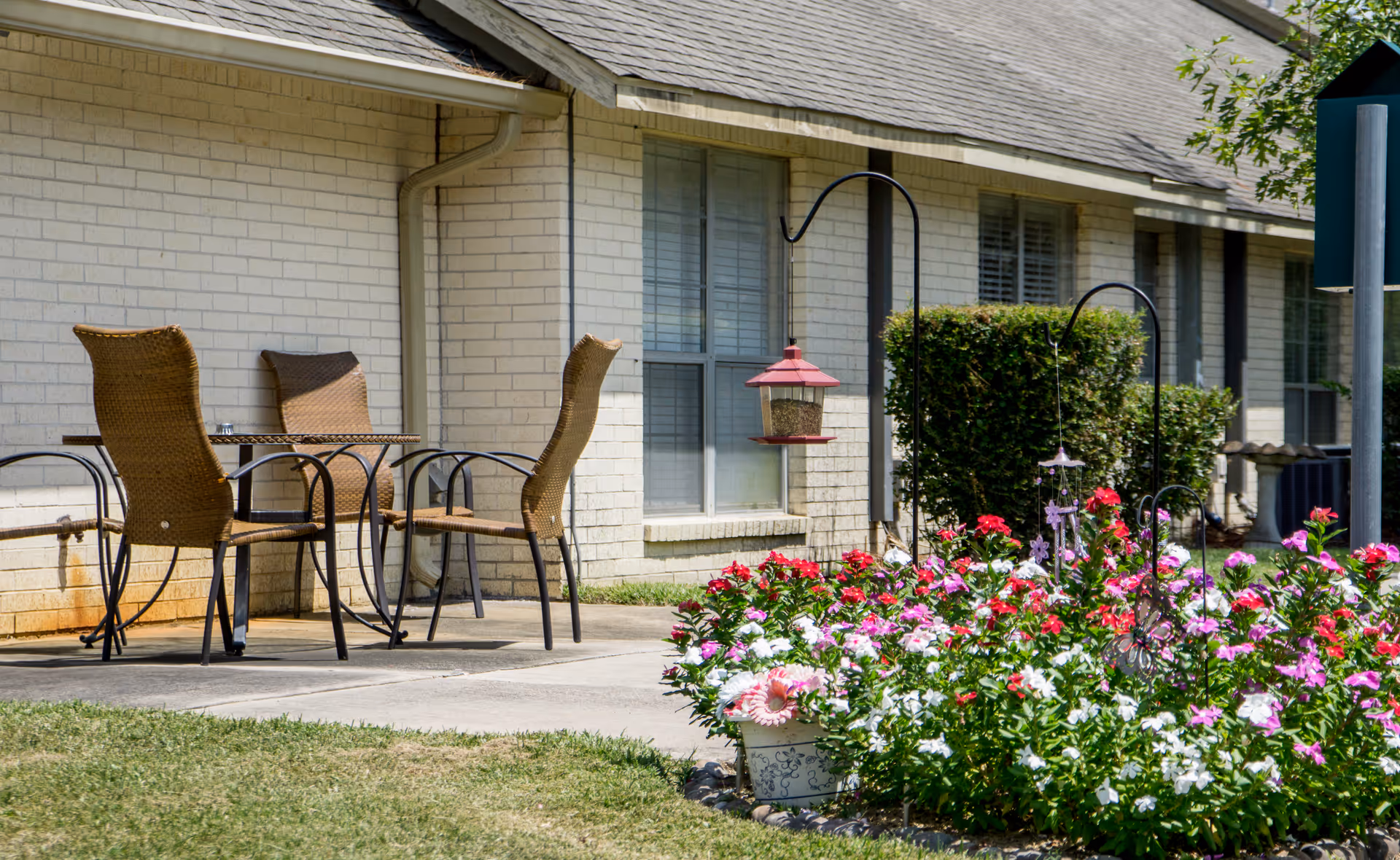 Patio with wicker chairs and a table beside a single-story brick building, with colorful flowers and bird feeders in the foreground.