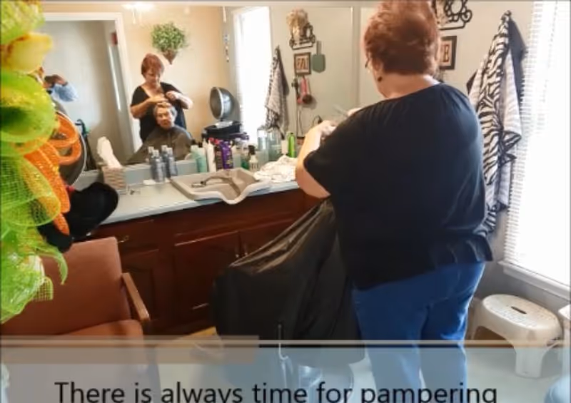 A woman is styling another woman's hair in a salon-like room with a large mirror, various hair care products on the counter, and a chair. The room has a window with blinds and a small white stool nearby.