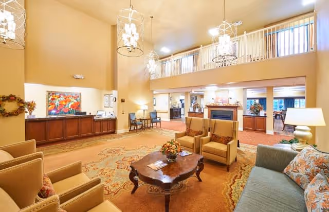 A spacious and well-lit senior living facility common area with high ceilings and hanging light fixtures. The room features comfortable seating including armchairs and a sofa around a wooden coffee table with a floral centerpiece. In the background, there is a reception desk, a fireplace, and additional seating areas. The decor includes warm tones, framed artwork, and floral arrangements.
