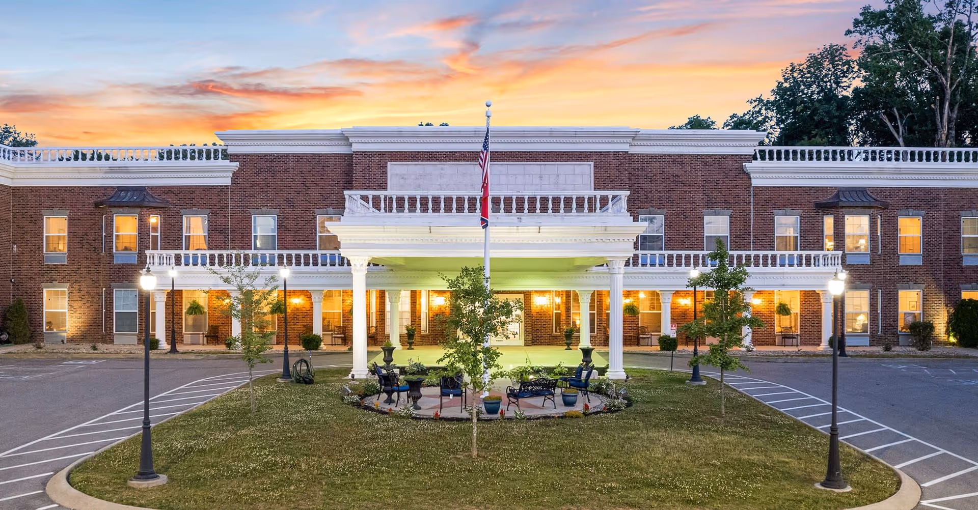 Front exterior view of Kempton of Hermitage facility at sunset, showing a large brick building with white columns and railings, a covered entrance with seating area, flagpole with American flag, and surrounding greenery and parking spaces.