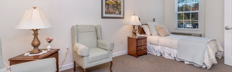 A cozy bedroom with a single bed covered in light-colored bedding and pillows, a wooden nightstand with a lamp and an alarm clock, a comfortable armchair, a wooden side table with a lamp and a small vase of flowers, a framed picture on the wall, and a window showing a view outside.