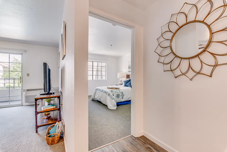 View of a hallway leading to a bedroom with a bed, bedside lamp, and window. To the left, there is a living room area with a TV on a stand, a sliding glass door, and a basket with books. A decorative round mirror with a sunburst frame hangs on the right wall.