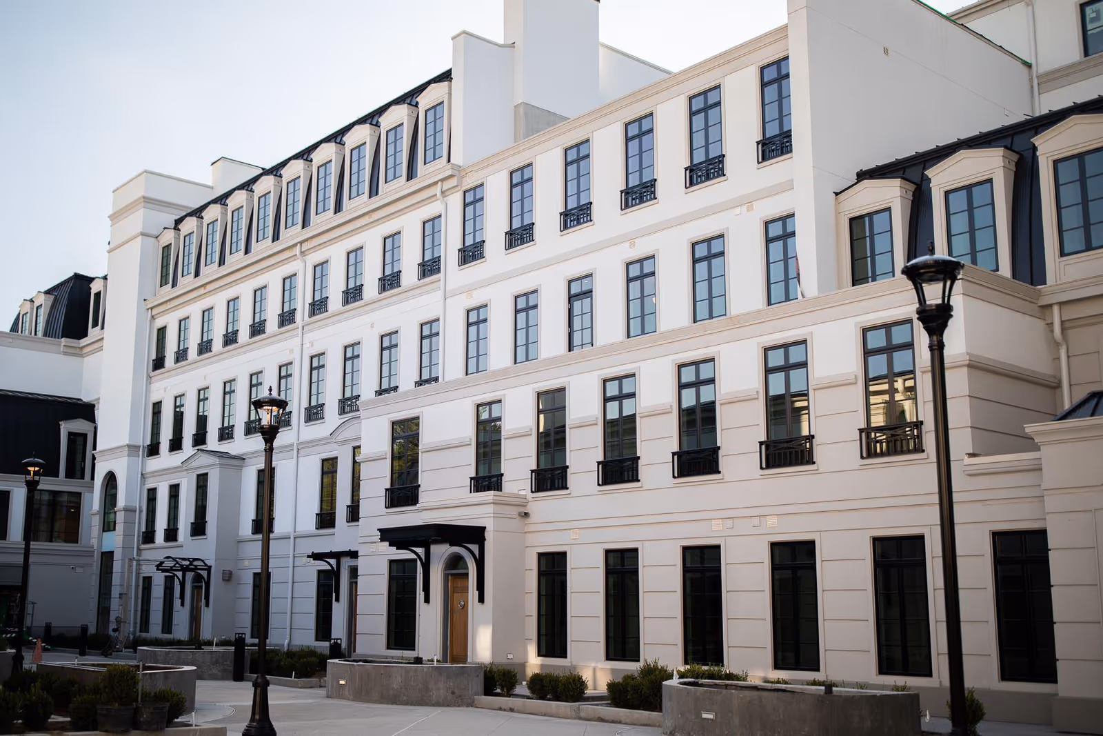 Exterior view of a multi-story white building with many windows and black trim, featuring street lamps and small landscaped areas in front.