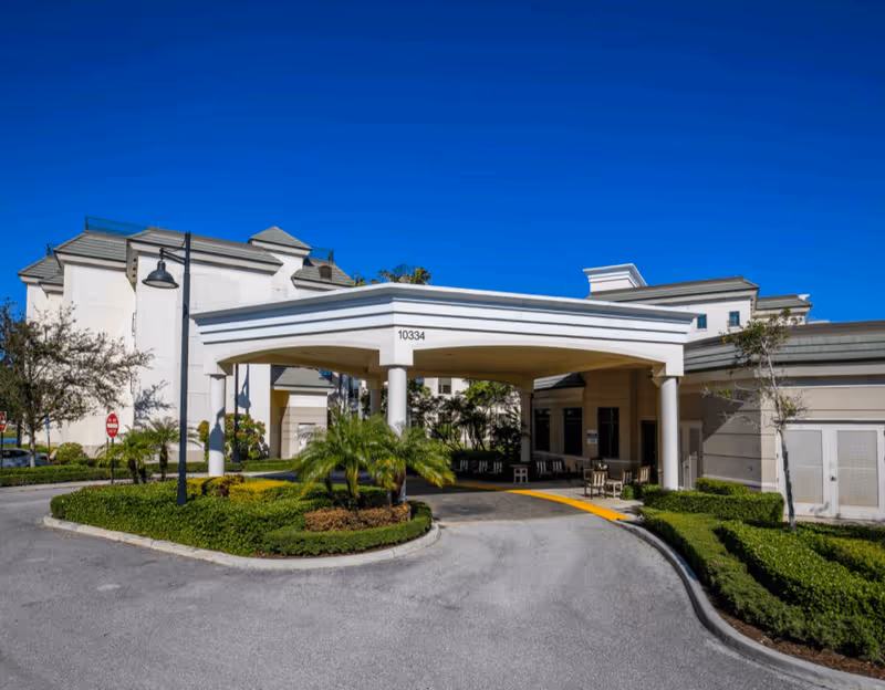 Exterior view of Luxe Senior Living at Wellington showing the main entrance with a covered drop-off area supported by white columns, surrounded by landscaped greenery and palm trees under a clear blue sky.