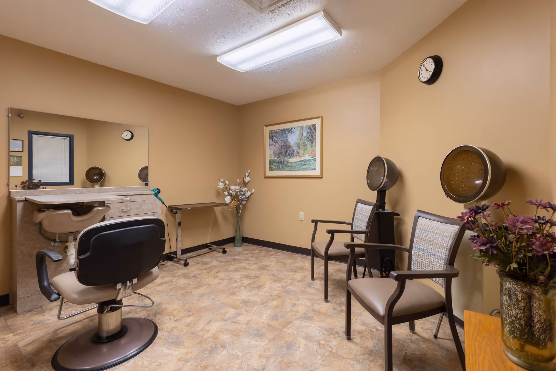 Interior view of a salon area in a senior living facility with beige walls and tiled floor. The room contains a salon chair facing a large mirror mounted on the wall, two hair drying stations with chairs, a small table with a vase of flowers, and a framed landscape painting on the wall. A clock is mounted on the wall above the hair drying stations.