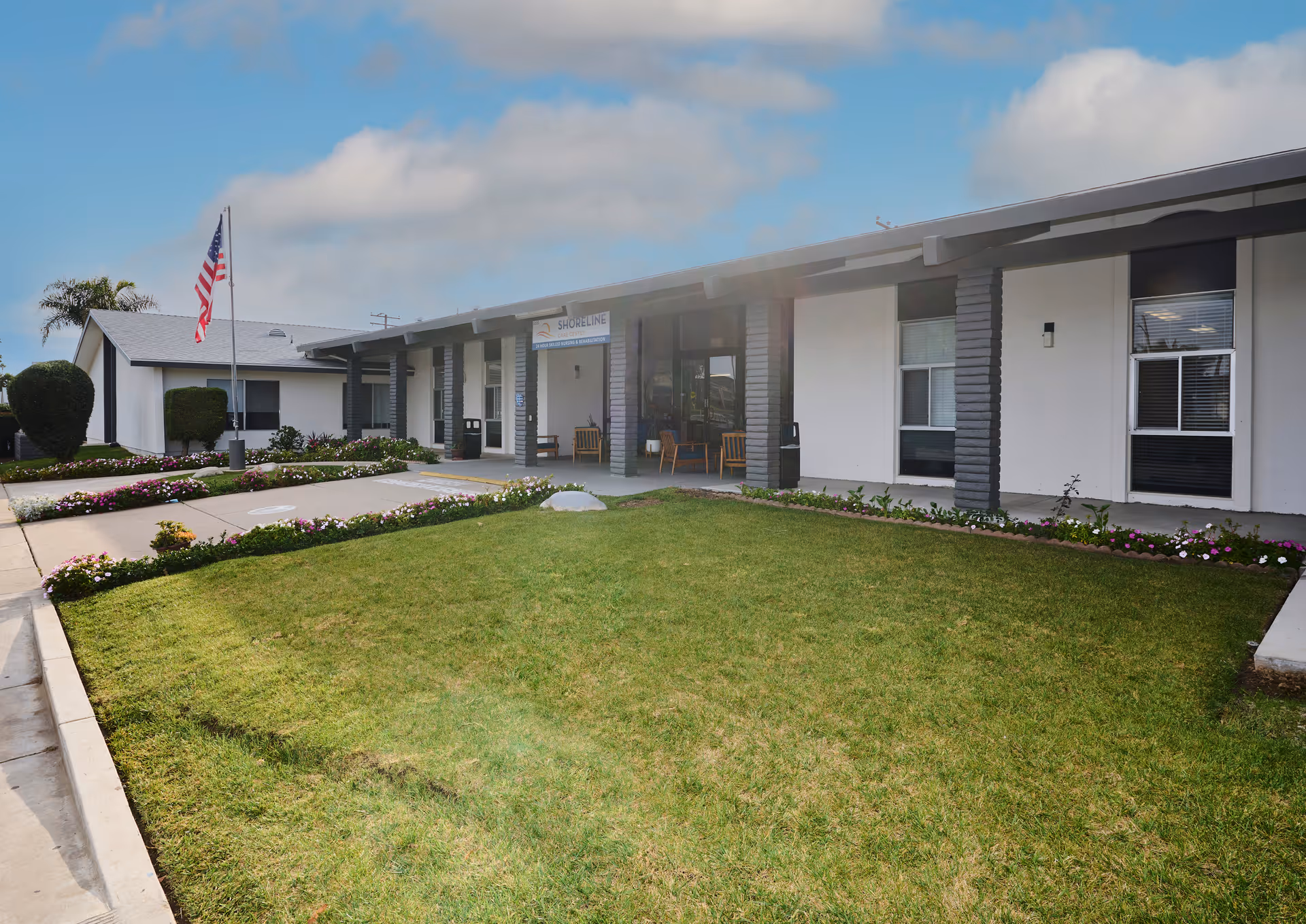 Front lawn and covered entrance of a single-story care facility with an American flag and chairs on the porch.