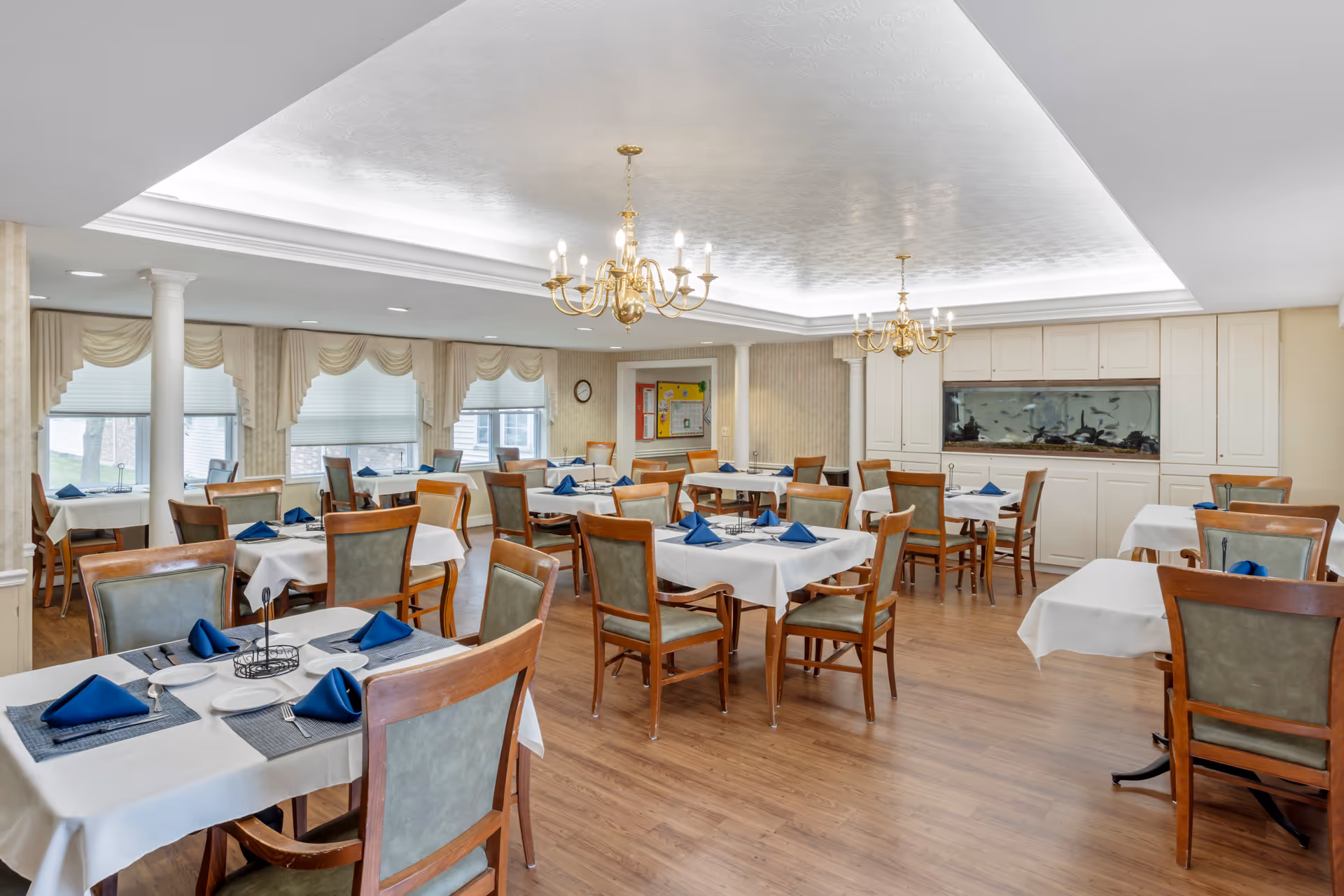 A dining room with multiple tables covered in white tablecloths and set with blue napkins, plates, and silverware. The room features wooden chairs with green upholstery, two gold chandeliers hanging from a white ceiling, large windows with beige curtains, and a built-in aquarium on the far wall.