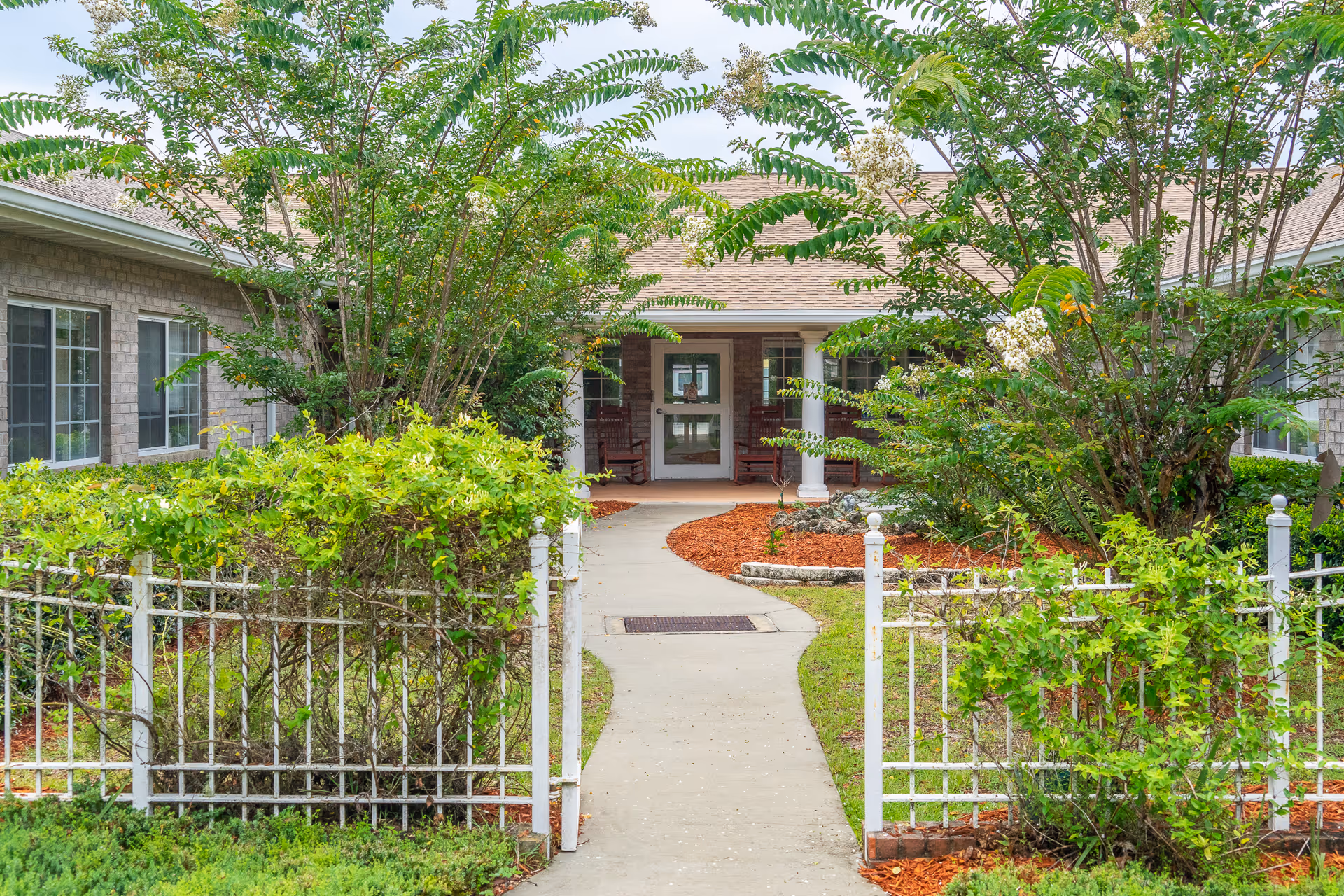 Entrance pathway leading to the front door of a nursing home building, surrounded by green bushes, trees, and a white metal fence on both sides.