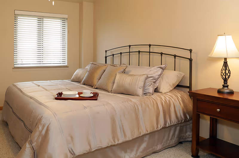 A neatly made bed with a metal headboard in a bedroom. The bed has a beige comforter and several matching pillows. A wooden nightstand with a decorative lamp is beside the bed. A window with closed blinds is on the wall behind the bed.