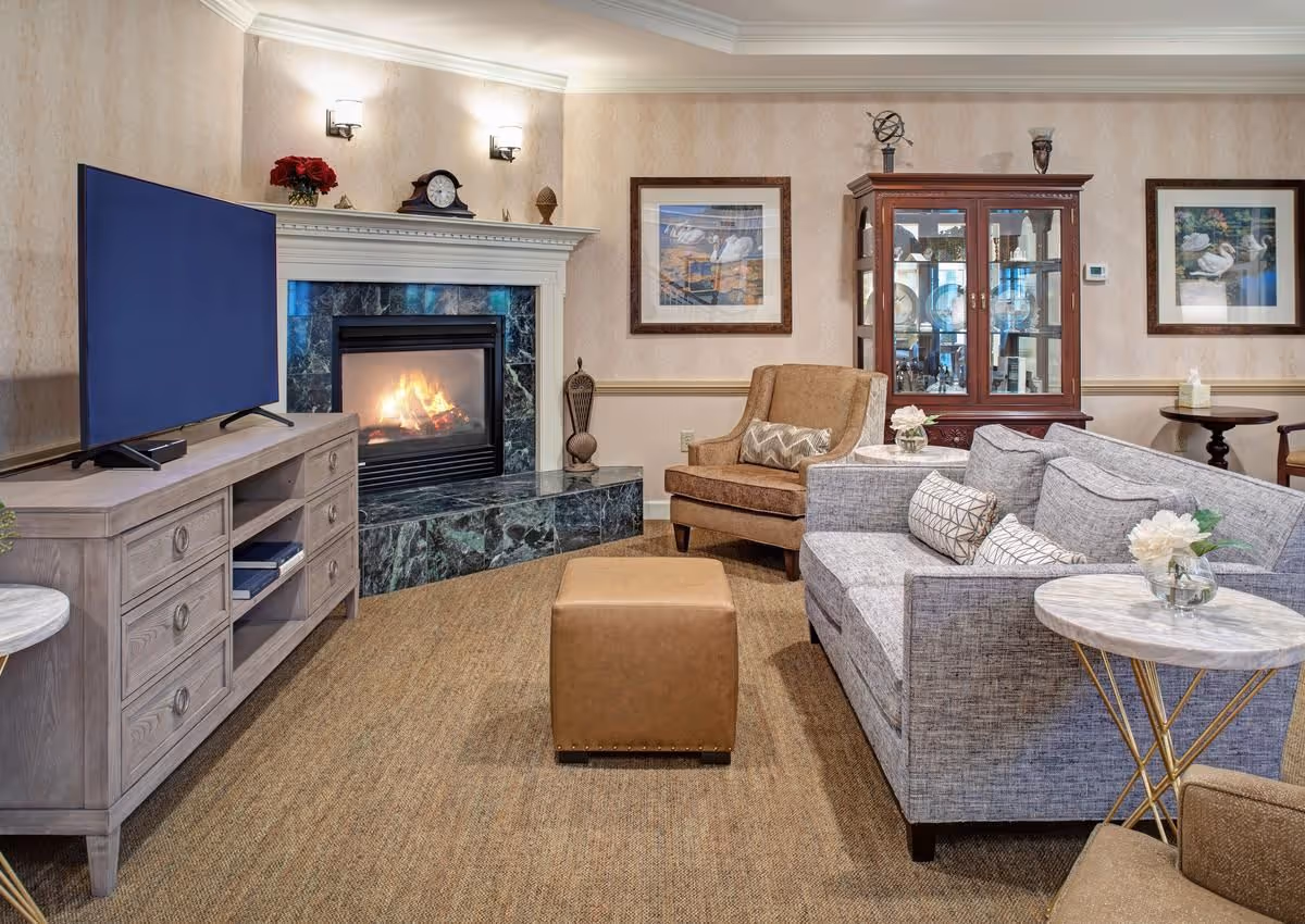Cozy common living room with a fireplace, television, sofa, armchairs, ottoman, and display cabinet.