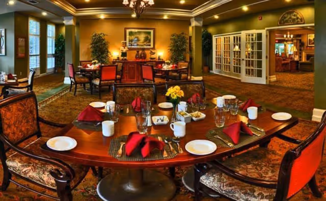 A warmly lit dining room with a large wooden table set for six people. The table is arranged with white plates, glasses, cups, silverware, and red folded napkins. The chairs around the table have patterned upholstery. In the background, there are more tables and chairs, decorative plants, a sideboard with vases, and framed artwork on the wall. The room has a carpeted floor and green walls with white trim.