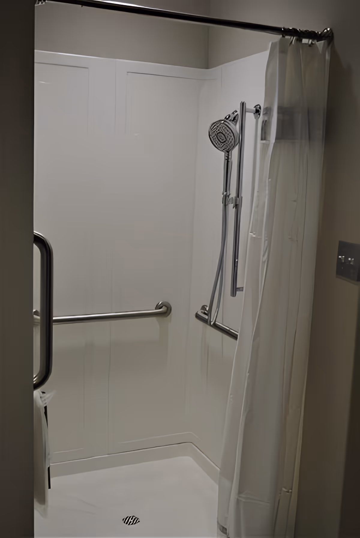 A clean, accessible shower stall with white walls, a handheld showerhead mounted on a vertical sliding bar, a white shower curtain partially drawn to the side, and stainless steel grab bars on the walls for support.
