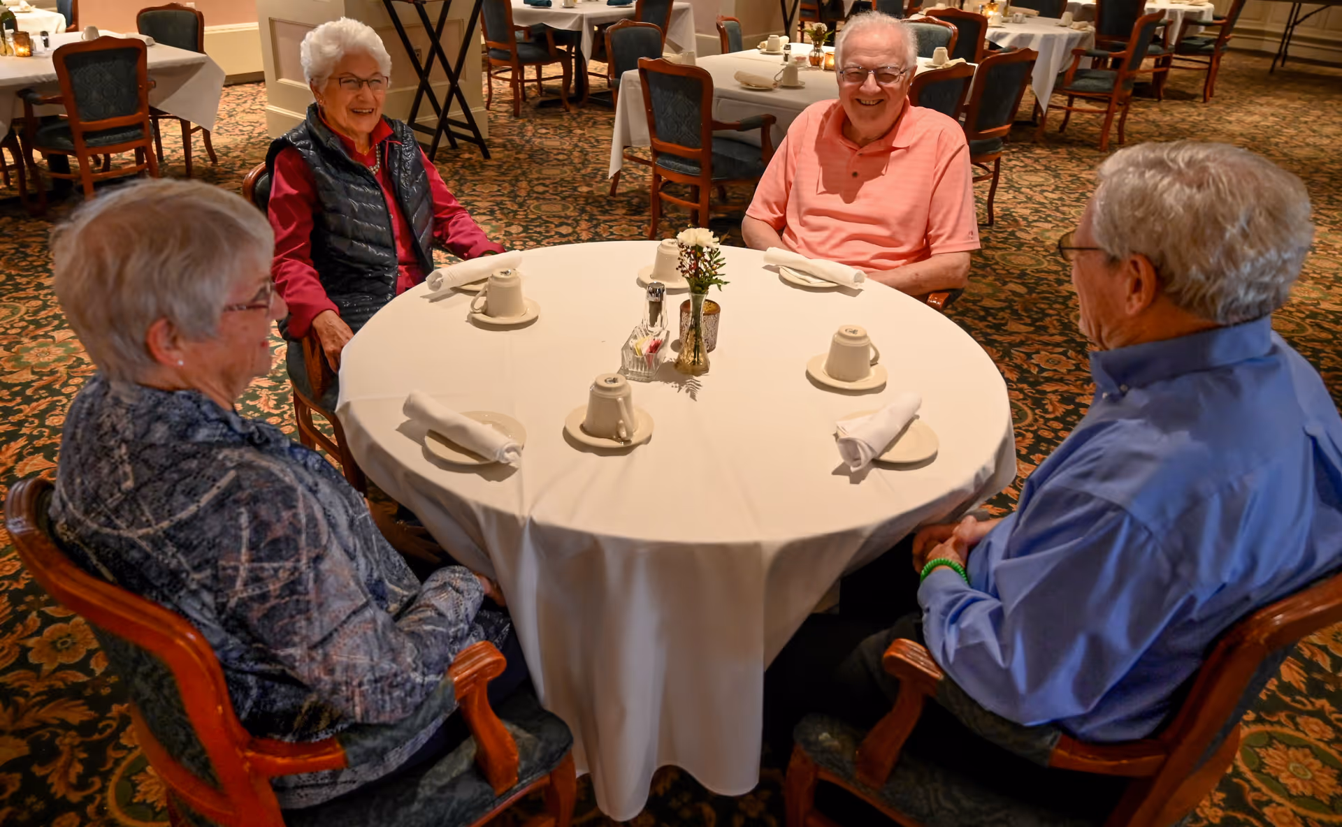 Four elderly residents seated around a round dining table set with cups and napkins in a dining room.