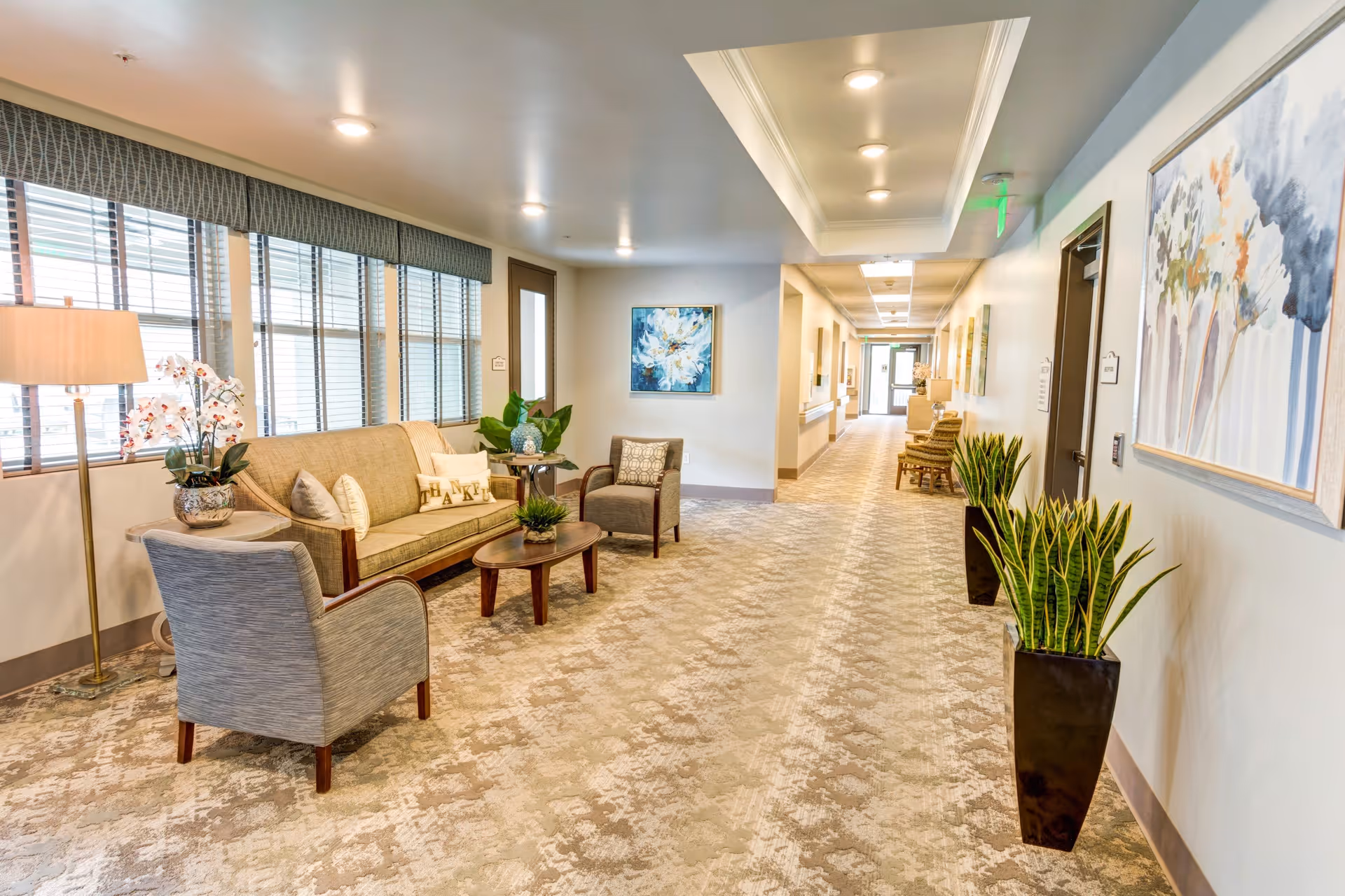 A bright and spacious hallway in a senior living facility featuring a seating area with a beige sofa, two armchairs, a coffee table, and decorative plants. The hallway is carpeted with a patterned design and has several framed artworks on the walls. Large windows with blinds allow natural light to fill the space.