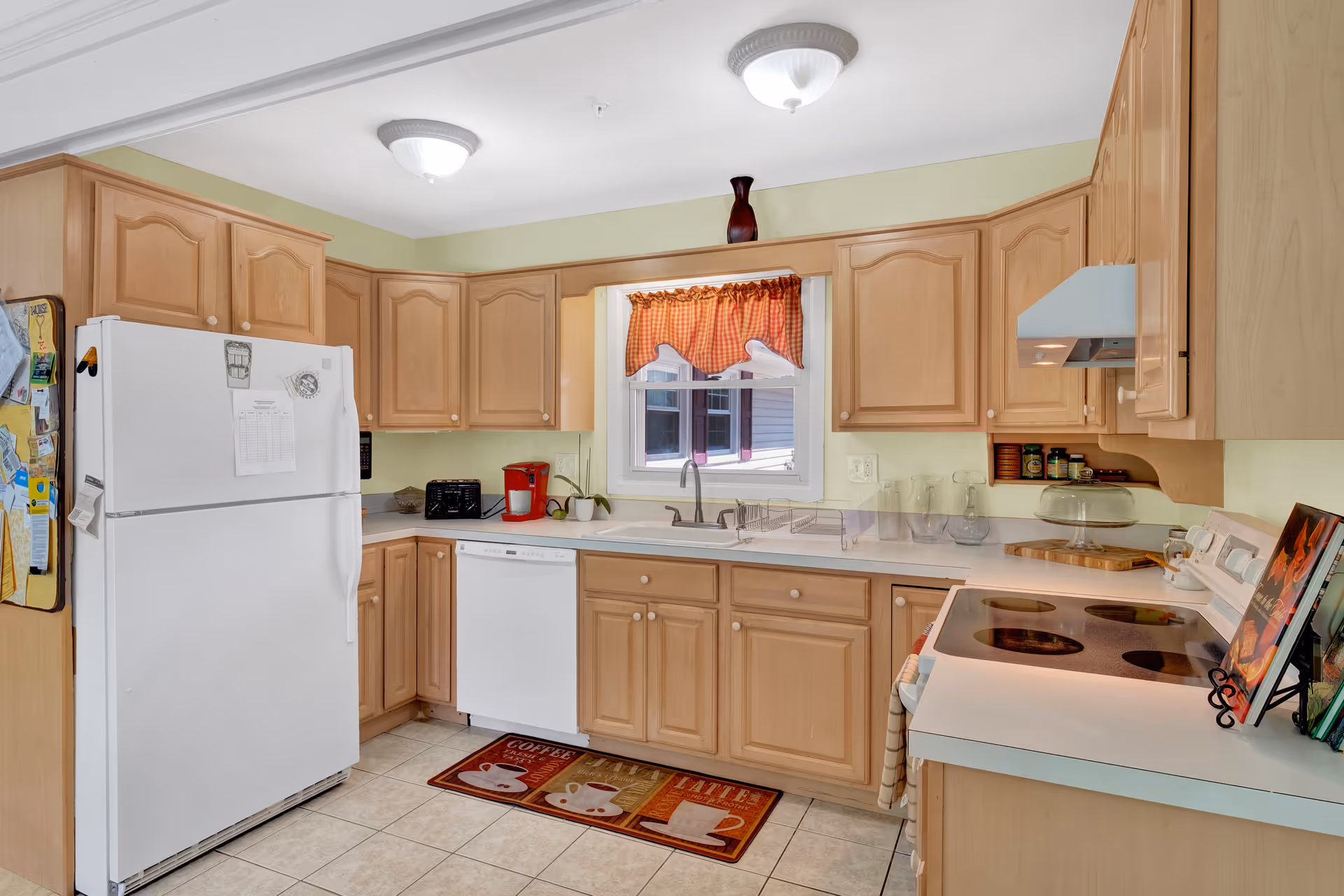 A kitchen with light wood cabinets, a white refrigerator, dishwasher, and stove. There is a window above the sink with a red and white checkered valance. Various kitchen items including a red coffee maker, toaster, and cookbooks are on the countertops. The floor is tiled and there is a colorful rug with coffee cup designs in front of the sink.
