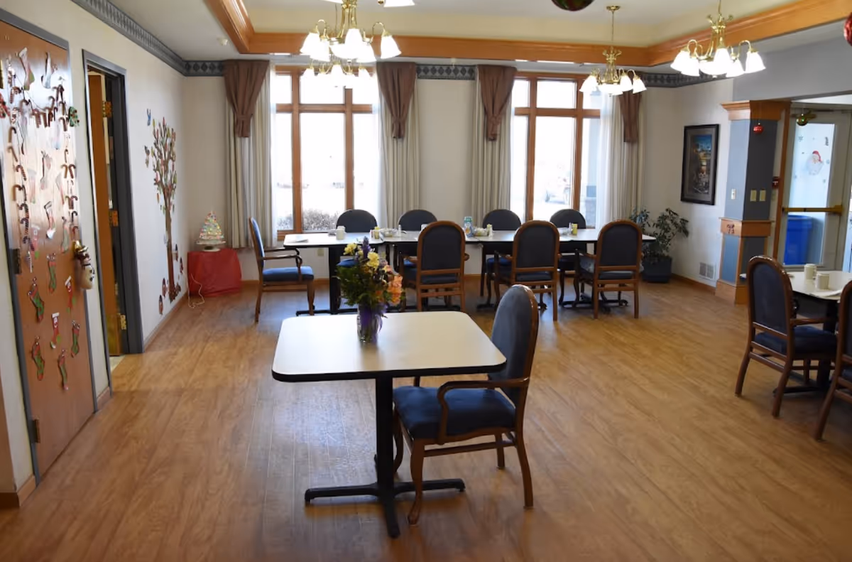 A dining room in an assisted living facility with wooden floors, several tables and chairs, large windows with curtains, and ceiling light fixtures. The room is decorated with holiday-themed items on the door and wall, and a vase with flowers is placed on the table in the foreground.