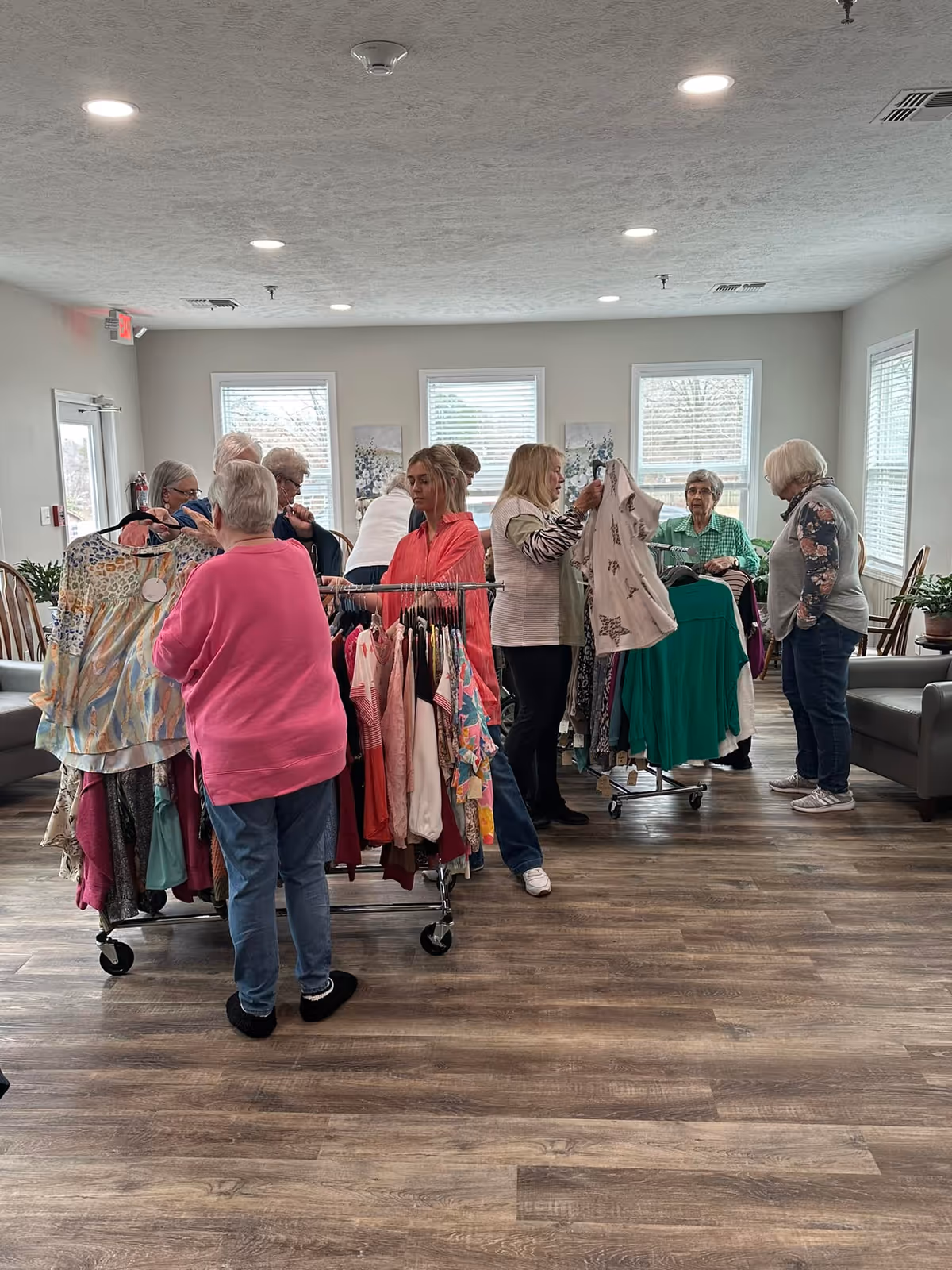 A group of elderly women browsing and examining clothing on racks inside a well-lit room with wooden floors and large windows. The room has light-colored walls, ceiling lights, and some chairs and plants along the sides.