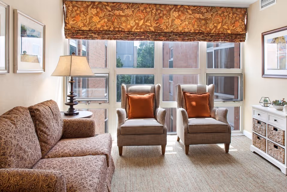 A cozy living room area with a patterned brown sofa on the left, a wooden side table with a lamp, two beige armchairs with orange cushions facing forward, large windows with a floral valance letting in natural light, and a white cabinet with wicker baskets and decorative items on the right.