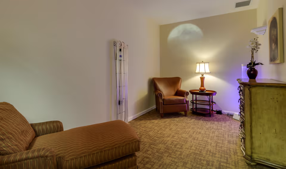 A cozy sitting area in a senior living facility featuring a brown upholstered chaise lounge on the left, a brown leather armchair next to a round wooden side table with a lit table lamp, a decorative chest of drawers on the right, and a wall-mounted painting above it. The room has beige walls and carpeted flooring.