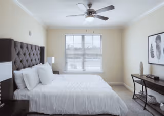 A bright bedroom with a large bed featuring a tufted dark headboard, white bedding, and multiple pillows. There is a nightstand with a lamp on the left side of the bed. A window with blinds is centered on the far wall, allowing natural light to fill the room. On the right side, there is a desk with a chair, decorative items, and framed artwork hanging above it. A ceiling fan with lights is mounted on the ceiling.