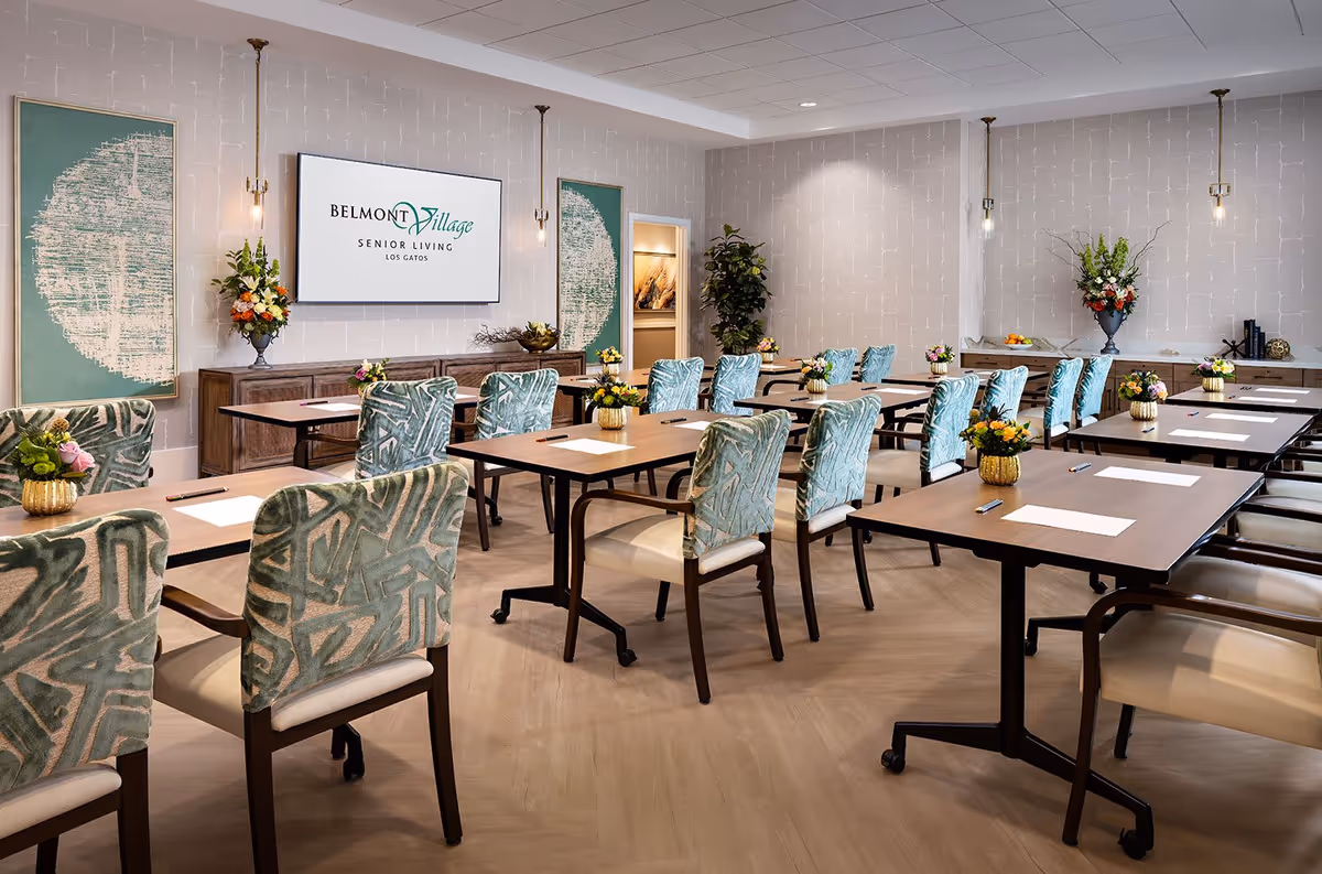 A well-lit meeting or conference room with multiple tables arranged in rows, each accompanied by patterned upholstered chairs. The tables have papers and pens placed on them, and small flower arrangements are set on each table. The room features light-colored walls with modern artwork and a large screen displaying the Belmont Village Senior Living Los Gatos logo. There are pendant lights hanging from the ceiling and decorative plants in the corners.