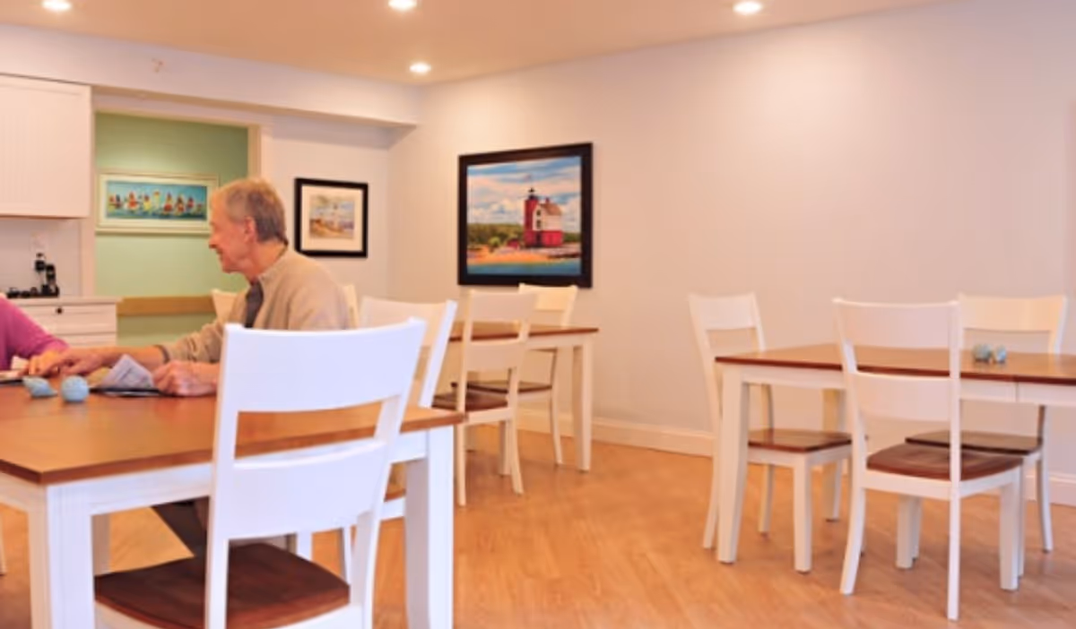 Bright dining room with white chairs and wooden tables; residents chat with a caregiver.