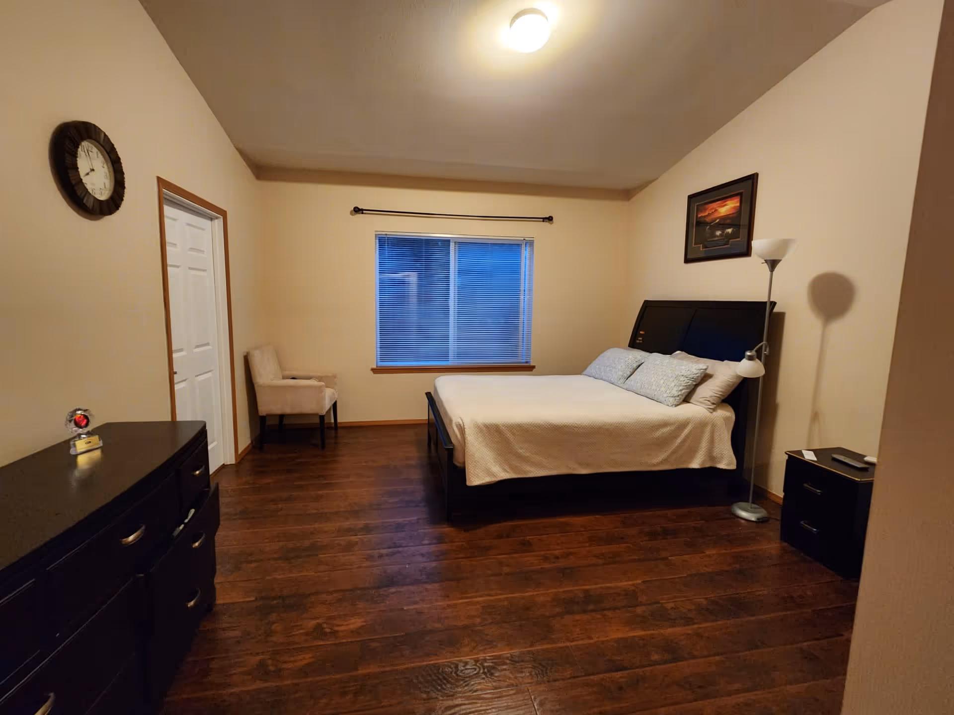 A bedroom with a large bed covered in a white bedspread and two pillows. There is a black headboard, a floor lamp, a small black nightstand with a phone on it, a beige upholstered chair near a window with closed blinds, a black dresser with drawers, and a wall clock. The room has wooden flooring and cream-colored walls with a framed picture above the bed.
