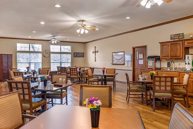 Bright communal dining room with multiple tables and chairs, ceiling fans, windows, and a small kitchenette.