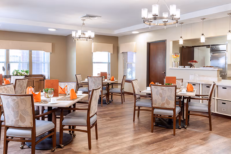 A bright dining room with several tables set with white cups, plates, and orange napkins. The room has wooden floors, beige walls, and multiple windows with beige valances. There are upholstered chairs with a leaf pattern around the tables. In the background, there is a counter area with a beverage dispenser and a door leading to another room.