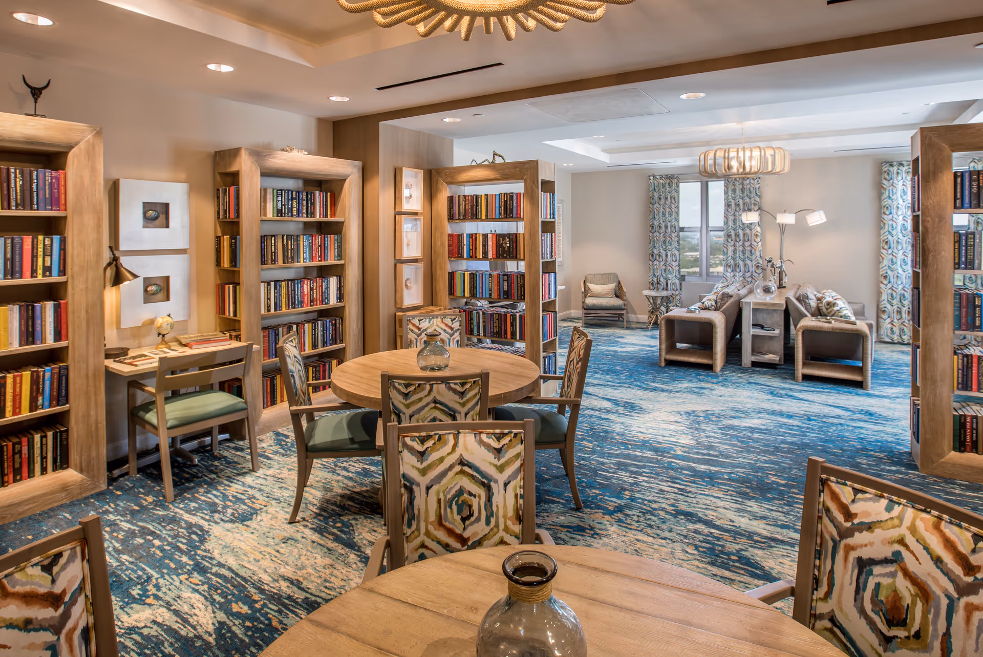 A cozy senior living facility common area featuring multiple wooden bookshelves filled with books, round wooden tables with patterned chairs, and a seating area with sofas and armchairs near windows with patterned curtains. The room has a blue and beige patterned carpet and warm lighting fixtures.