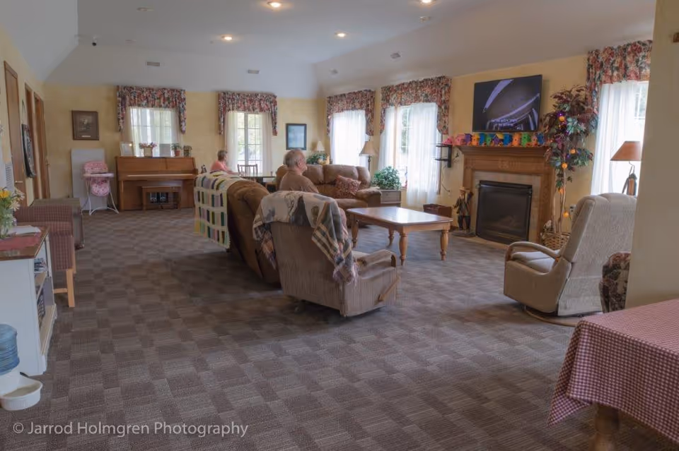 Communal living room with sofas, armchairs, a coffee table and fireplace, windows with floral valances and a few seated people.