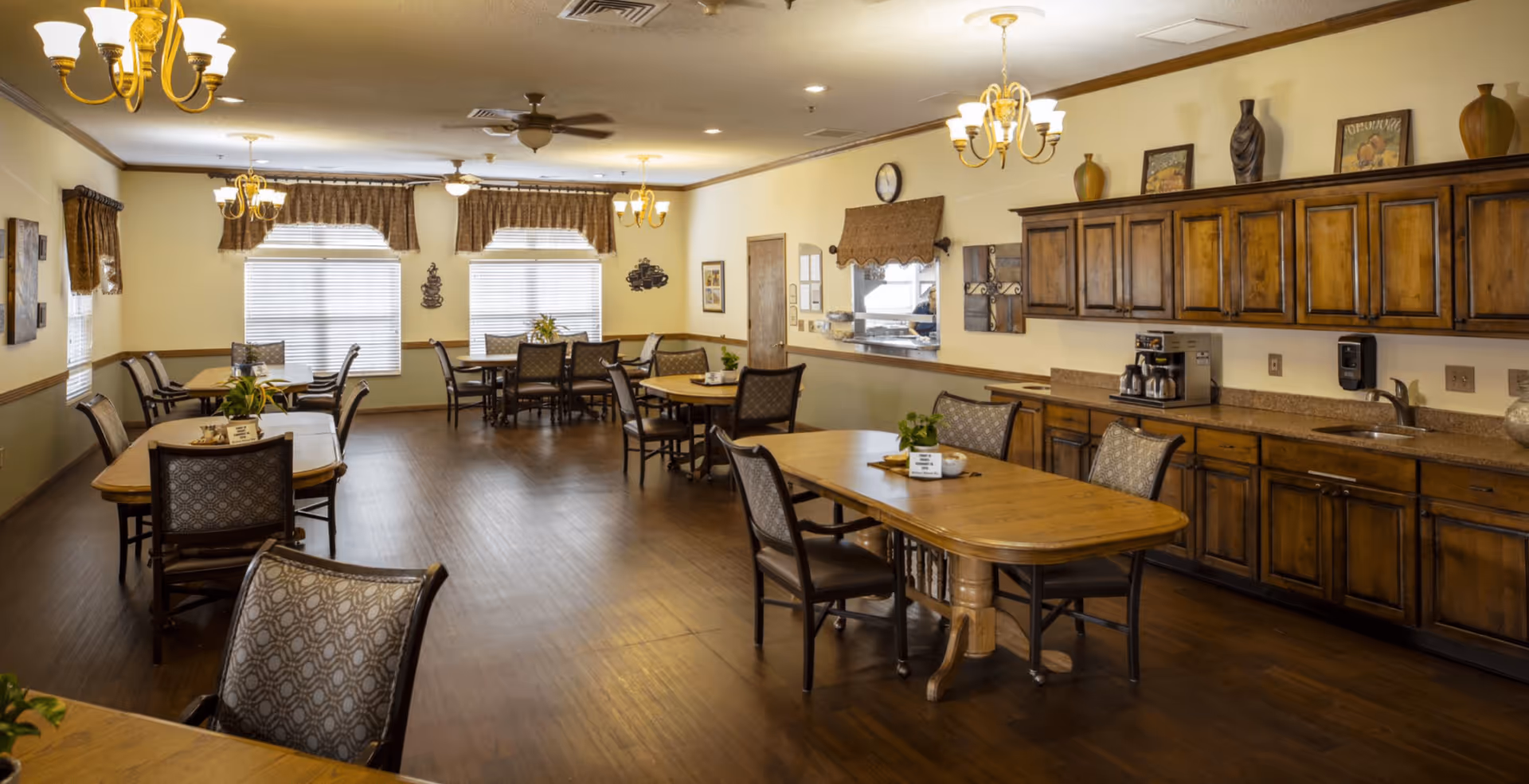 A spacious dining room with several wooden tables and cushioned chairs arranged neatly. The room features wooden flooring, warm yellow walls with decorative trim, and large windows with valances letting in natural light. There are chandeliers and ceiling fans on the ceiling, and a long counter with wooden cabinets, a coffee machine, and a sink along one wall.