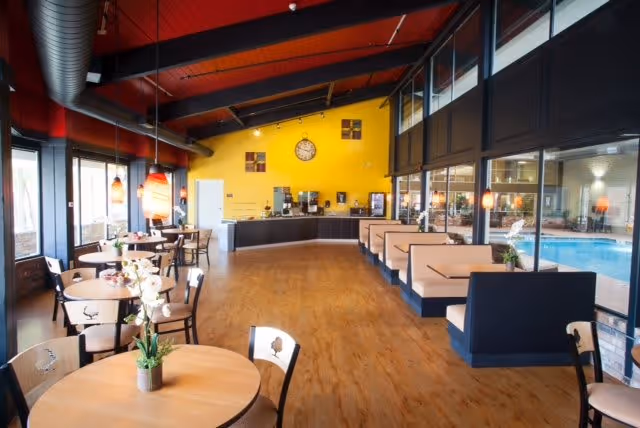 A bright and spacious dining area with wooden floors, round tables with chairs, and booth seating along a glass wall overlooking an indoor pool. The ceiling is red with exposed ductwork, and the far wall is painted yellow with a large clock and a coffee station underneath.