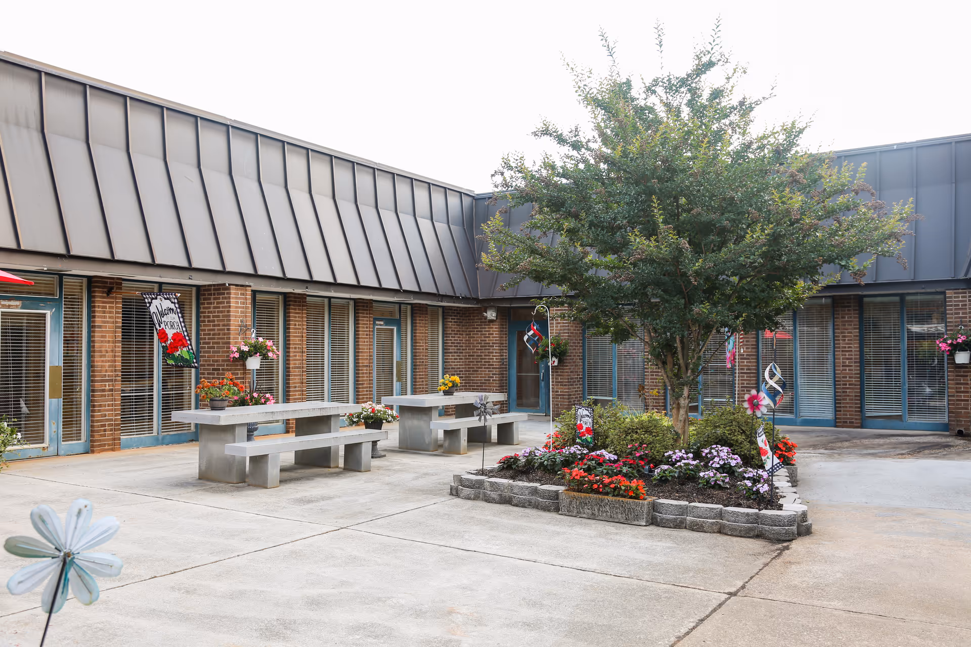 Outdoor courtyard with concrete picnic tables, a central flower bed and tree, and surrounding brick building facades with windows and doors.