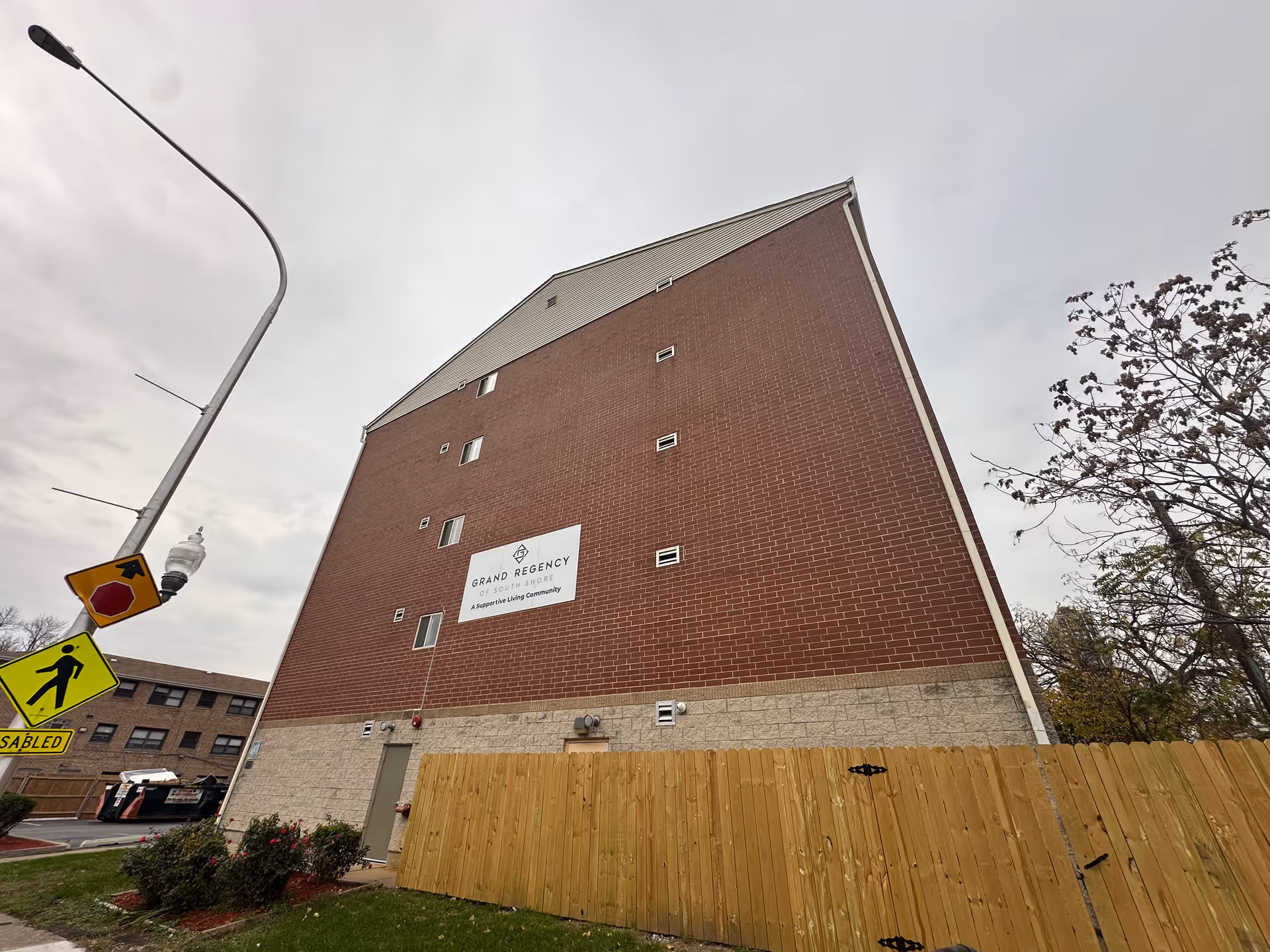 Tall brick apartment-style building with a 'Grand Regency' sign, a wooden fence, and street signs in the foreground.
