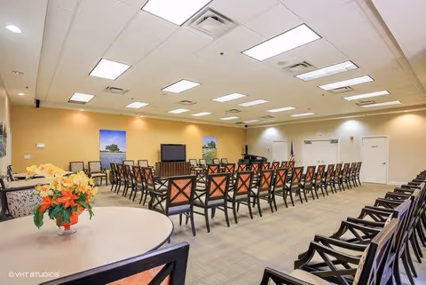 A large meeting or activity room with rows of chairs arranged in a U-shape facing a television and a piano in the corner. The room has beige walls, a carpeted floor, and fluorescent ceiling lights. There are two landscape paintings on the far wall and a round table with a flower arrangement in the foreground.