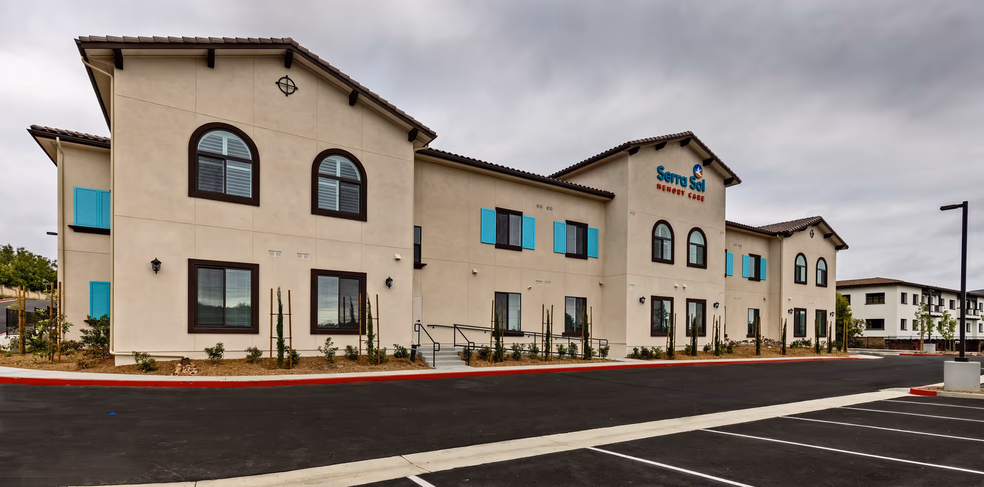 Exterior view of Serra Sol Memory Care building with beige walls, brown trim, and turquoise window shutters. The building has two stories with arched windows and a tiled roof. There is a parking lot in front and a cloudy sky overhead.